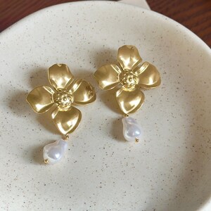 May include: A pair of gold-toned metal earrings shaped like four-petaled flowers with textured centers. Each earring features a single white, irregular-shaped drop pearl suspended from the bottom petal, creating a delicate and balanced appearance.