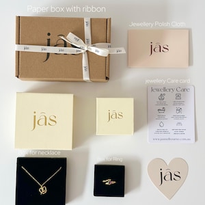 May include: An assortment of jewelry packaging, including a brown paper box with a white ribbon, cream-colored boxes, and black velvet boxes. A gold necklace and ring are displayed. The packaging includes a jewelry care card and a polishing cloth.