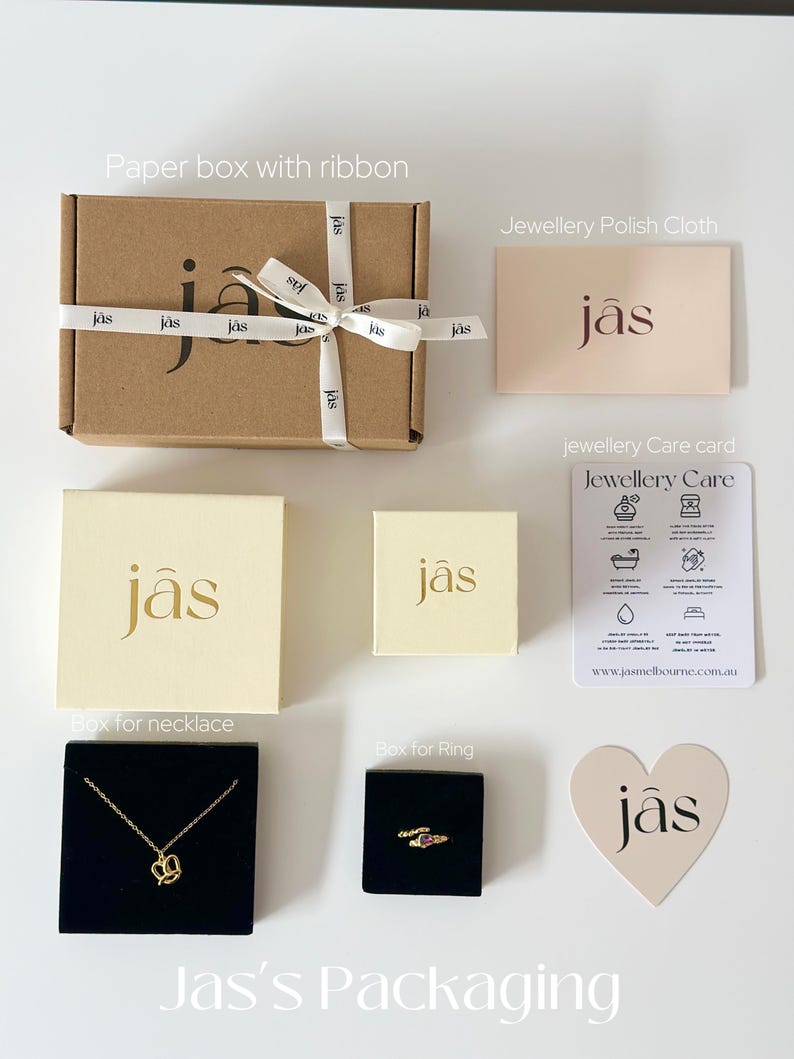 May include: An assortment of jewelry packaging, including a brown paper box with a white ribbon, cream-colored boxes, and black velvet boxes. A gold necklace and ring are displayed. The packaging includes a jewelry care card and a polishing cloth.