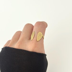 May include: A gold-colored ring in the shape of angel wings. The ring features detailed feather textures and is worn on a finger. The background is plain white, highlighting the ring's design.