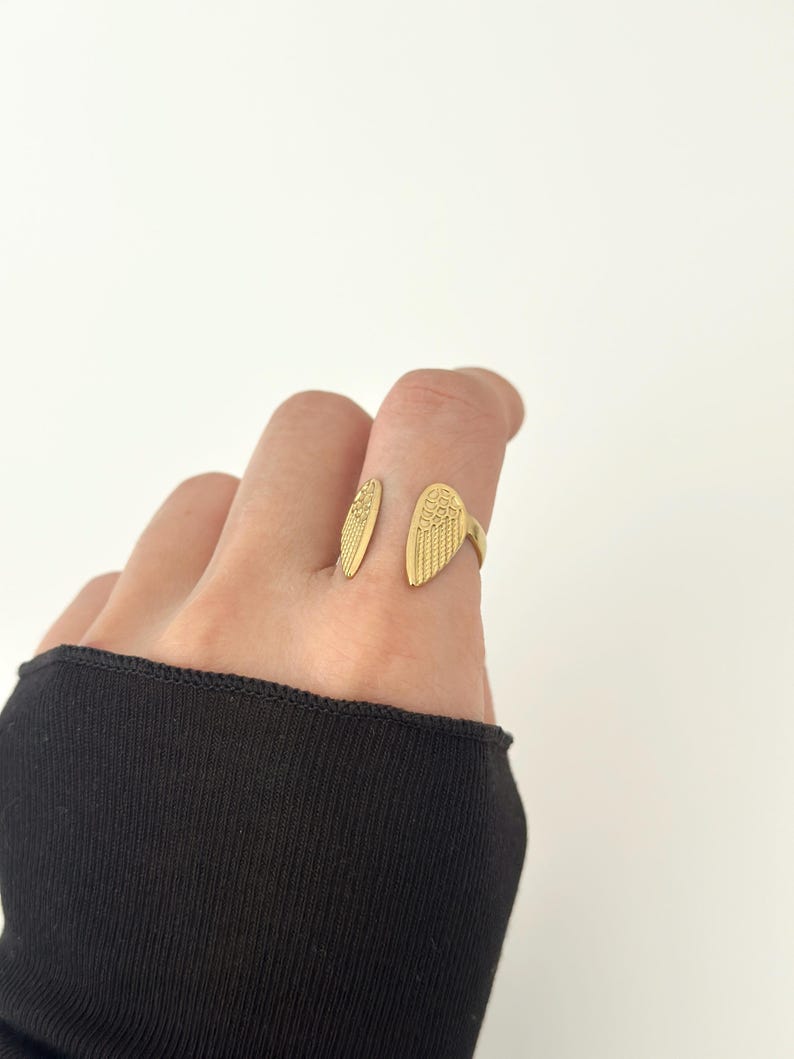 May include: A gold-colored ring in the shape of angel wings. The ring features detailed feather textures and is worn on a finger. The background is plain white, highlighting the ring's design.