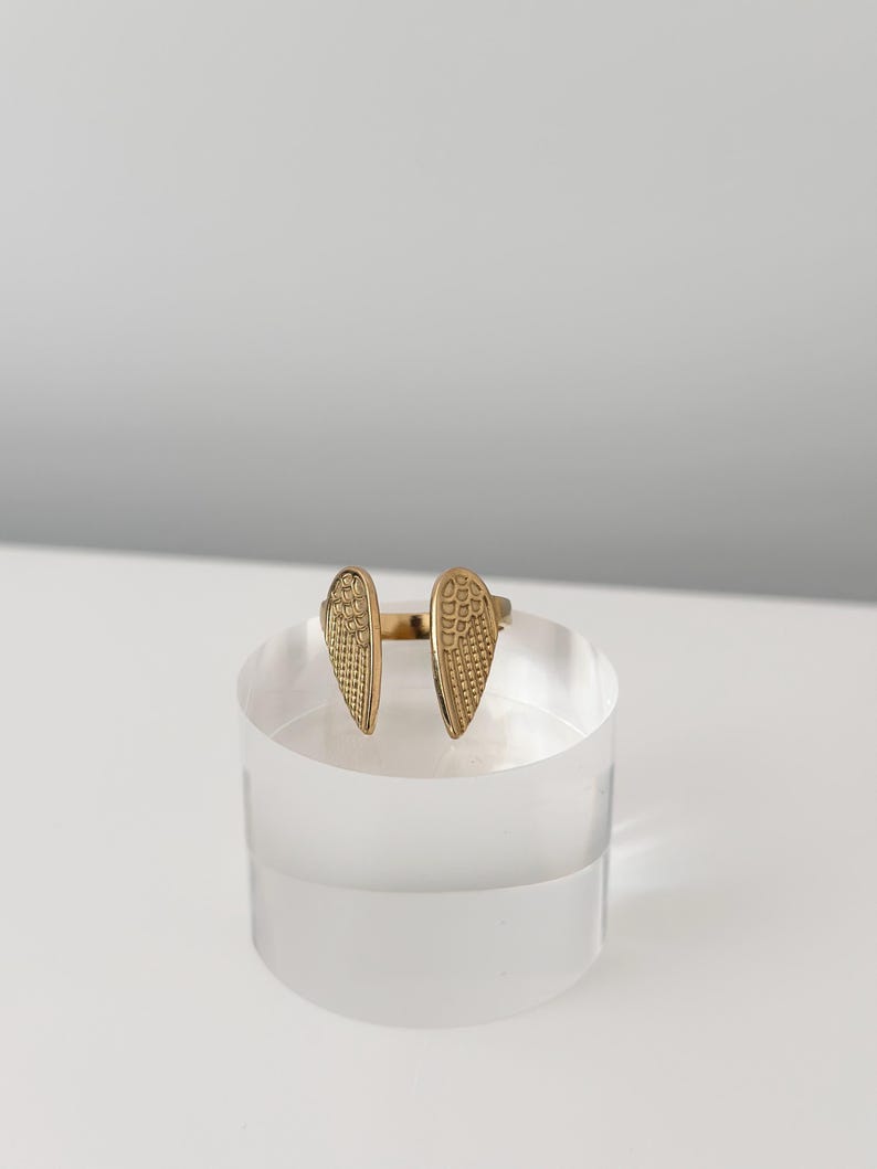 May include: A gold-coloured ring with an open design, featuring two stylised wing shapes. Each wing has a textured surface with a repeating pattern. The ring is displayed on a clear, cylindrical stand against a white background.