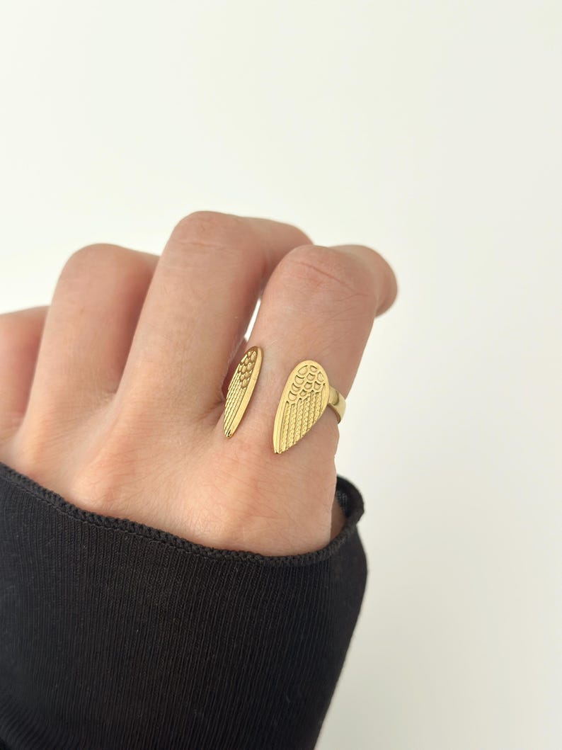 May include: A gold-colored ring with an open design, featuring wing-shaped details. The ring is worn on a finger, with the wings extending on either side. The ring's surface has a textured pattern, and the overall aesthetic is elegant and symbolic.