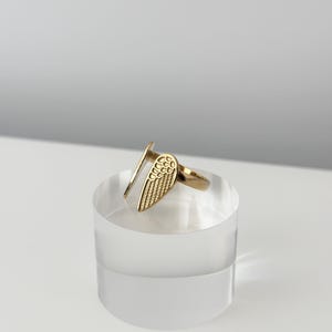 May include: A gold-colored ring featuring a stylized wing design. The ring is open-ended and sits on a clear, cylindrical display stand. The wing detail is textured, and the ring appears to be made of metal.
