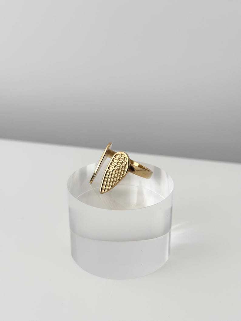 May include: A gold-colored ring featuring a stylized wing design. The ring is open-ended and sits on a clear, cylindrical display stand. The wing detail is textured, and the ring appears to be made of metal.