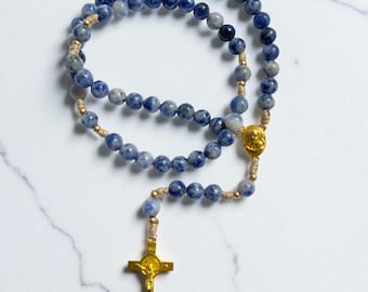 May include: A rosary with blue and white speckled beads. The beads are strung on a beige cord and connected with gold-colored metal pieces, including a crucifix and a centerpiece.