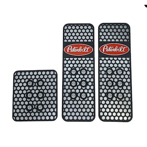 May include: Three black metal pedal pads with a honeycomb pattern and the Peterbilt logo in red. The pads are designed to be used in a truck cab.