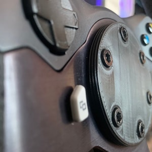 Puede incluir: Primer plano de un volante negro con un bot&oacute;n plateado y una placa redonda negra con seis tornillos. La placa tiene una superficie texturizada.