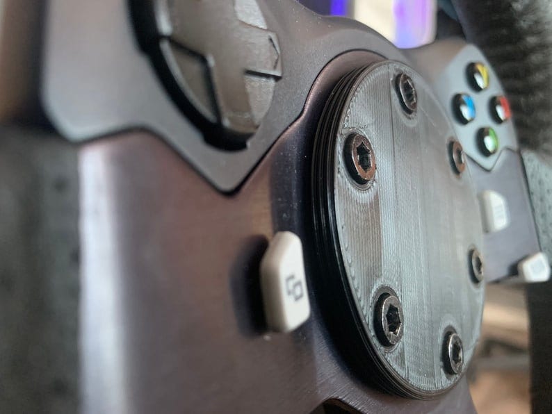 Puede incluir: Primer plano de un volante negro con un bot&oacute;n plateado y una placa redonda negra con seis tornillos. La placa tiene una superficie texturizada.