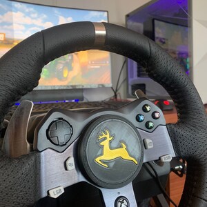 Puede incluir: Un volante de carreras negro con un logotipo amarillo y verde de un ciervo. El volante tiene botones y paletas para jugar.