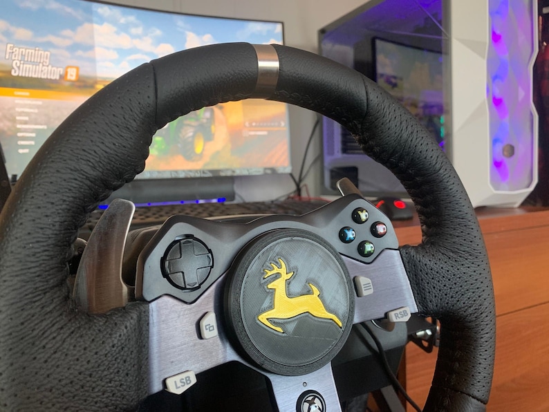 Puede incluir: Un volante de carreras negro con un logotipo amarillo y verde de un ciervo. El volante tiene botones y paletas para jugar.