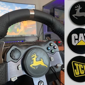 Puede incluir: Un volante de carreras negro con un borde plateado y un logotipo de ciervo amarillo y negro en el bot&oacute;n central. El volante est&aacute; conectado a una consola de juegos con botones y una palanca de mando. El fondo muestra una pantalla de ordenador con un videojuego.