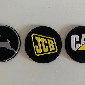 Puede incluir: Tres parches redondos negros con diferentes logotipos. El primer parche tiene una silueta de ciervo plateada sobre un fondo negro. El segundo parche tiene un logotipo JCB amarillo sobre un fondo negro. El tercer parche tiene un logotipo CAT blanco con un tri&aacute;ngulo amarillo sobre un fondo negro.