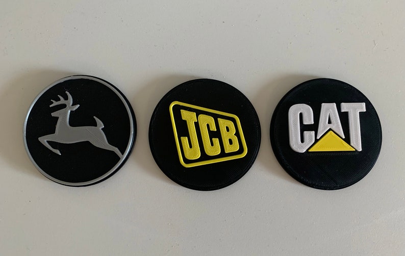 Puede incluir: Tres parches redondos negros con diferentes logotipos. El primer parche tiene una silueta de ciervo plateada sobre un fondo negro. El segundo parche tiene un logotipo JCB amarillo sobre un fondo negro. El tercer parche tiene un logotipo CAT blanco con un tri&aacute;ngulo amarillo sobre un fondo negro.