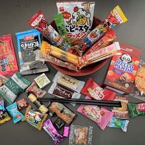 May include: A selection of Japanese snacks, including Hello Panda biscuits, Kit Kat, and various packaged sweets and crackers. The snacks are arranged on a red plate and a dark surface.