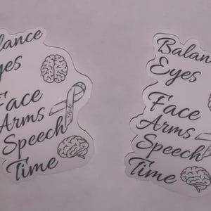 Peut inclure: Deux autocollants blancs avec du texte et des illustrations noirs. Le texte indique "Balance Eyes Face Arms Speech Time" avec des illustrations de cerveau et de ruban. Les autocollants sont sur un fond gris clair.