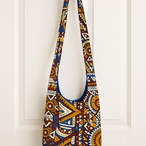 May include: A shoulder bag with a colorful geometric pattern in shades of brown, yellow, blue, and white. The bag has a long strap and a blue interior. The bag is hanging on a white door.