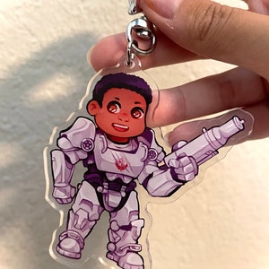 May include: A white and purple cartoon character wearing a white and gray power armor suit and holding a gun. The character has brown skin and a purple afro. The character is a keychain.