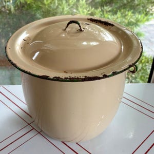 May include: A cream-colored enamel chamber pot with a matching lid. The container features a dark green rim and a small handle on the lid, showing visible signs of rust and wear along the edges of the rim and the handle of the lid.