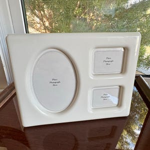 May include: A white ceramic picture frame with an oval and two rectangular openings. Each opening has the text "Place Photograph Here". The frame is on a dark brown surface.