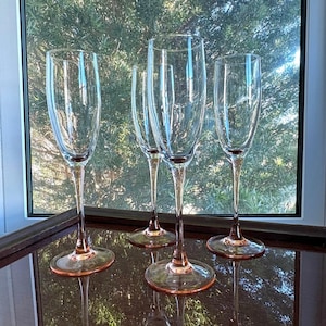 May include: Four clear champagne flutes with pink-tinted bases and stems. The glasses are tall and slender, reflecting light. The flutes are arranged on a reflective surface, with a window and greenery in the background.