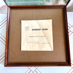 May include: A square wooden picture frame with a dark brown finish and a flat brown backing board. Centered on the backing is a paper insert labeled Sudberry House Needlework Picture Frame with instructions to frame your needlework as desired.