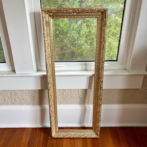 May include: A tall, rectangular picture frame with a metallic gold finish. The frame features an intricate, raised relief pattern along its entire border, consisting of repeating floral and scrollwork motifs that create a textured appearance.