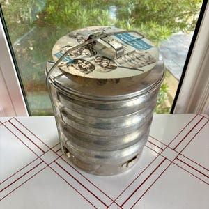 May include: A cylindrical, multi-tiered aluminum picnic carrier with a metal locking handle. A circular paper label on the lid features illustrations of food and the text "Picnic Pack by Buckeye." The stackable container has a metallic finish.