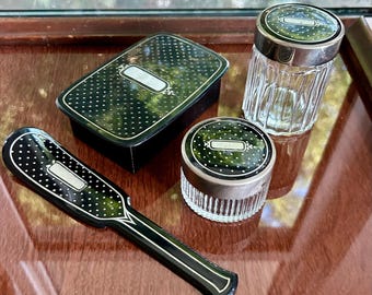 May include: A collection of vintage vanity items, including a black and white dotted brush, a small box, and two glass jars with silver-toned lids. The items are arranged on a wooden surface, showcasing a classic aesthetic.