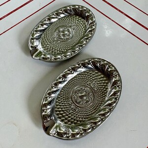 May include: Two oval-shaped silver-toned metal trays featuring a reflective finish. Each tray has a central circular medallion with floral detailing, surrounded by a textured pattern of small raised dots and a decorative scalloped border edge.