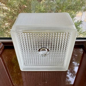 May include: A clear, square-shaped light fixture with a textured, ribbed top and a patterned, grid-like bottom. The fixture has a central circular element and is likely for indoor use. The fixture is made of glass.