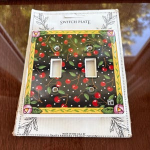 May include: A decorative double switch plate with a black background, adorned with red cherries and white polka dots. The plate has a yellow border with floral accents and the text "SWITCH PLATE". Made in the U.S.A. by Santa Barbara Ceramic Design.