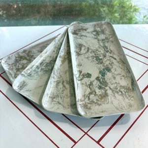May include: A set of three rectangular trays featuring a marbled pattern in shades of white, sage green, and muted gold. Each tray has slightly raised edges and a smooth, flat surface, arranged in a fan-like overlapping formation.