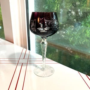 May include: A deep purple cut-to-clear crystal wine glass featuring intricate etched floral and grape cluster patterns on the bowl. The stem has a textured, faceted design leading down to a clear, circular base with etched decorative markings.