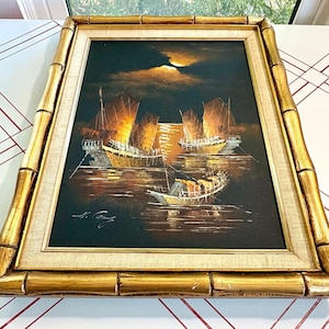 May include: An oil painting depicting several boats on a body of water at night, with a bright moon in the sky. The painting is framed in a gold bamboo-style frame.