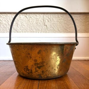 May include: A round, metallic pot with a weathered, golden-toned finish and a dark, arched handle attached to the sides. The exterior surface shows signs of oxidation and wear, featuring a wide, open top and a tapered base design.