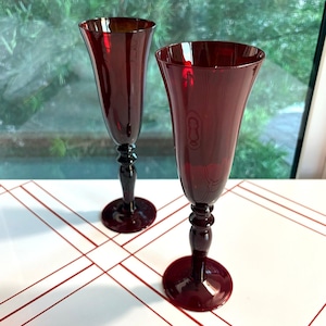 May include: Two tall, deep red glass champagne flutes with a smooth, flared bowl and a slender, multi-tiered stem leading to a circular base. The translucent ruby-colored glass catches the light, creating a rich, saturated appearance for each flute.
