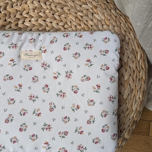 Peut inclure: Une housse d'ordinateur portable blanche à motifs floraux. Le motif présente de petits groupes de fleurs rouges, roses et jaunes avec des feuilles vertes. Une étiquette avec le texte "Maracuja PARIS" est cousue sur le devant.