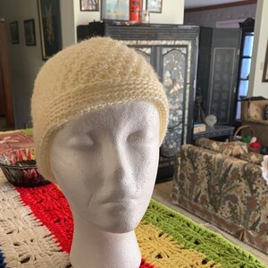 May include: A white crocheted beanie hat with a textured stitch pattern. The hat is displayed on a colorful crocheted blanket with stripes of blue, white, red, yellow, and green.