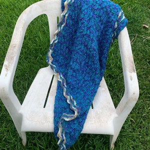 May include: A handmade, blue and teal crochet blanket with a scalloped edge in white, tan, and gray. The blanket is draped over a white plastic chair, set against a backdrop of green grass.