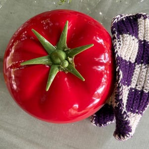 Könnte beinhalten: Ein roter Topf in Tomatenform mit einem grünen Plastikstiel und Blättern. Der Topf steht auf einer weißen Oberfläche mit einem lila-weißen Häkeltuch daneben.