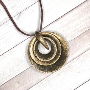 Puede incluir: Un collar dorado con un colgante que presenta tres círculos entrelazados. El colgante está unido a un cordón de cuero marrón.