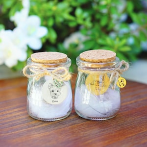 May include: Two small glass jars with cork lids, filled with white feathers and personalized with dog-themed labels. The jars are tied with twine and have a small gold charm with the word "LOVE" and a paw print.