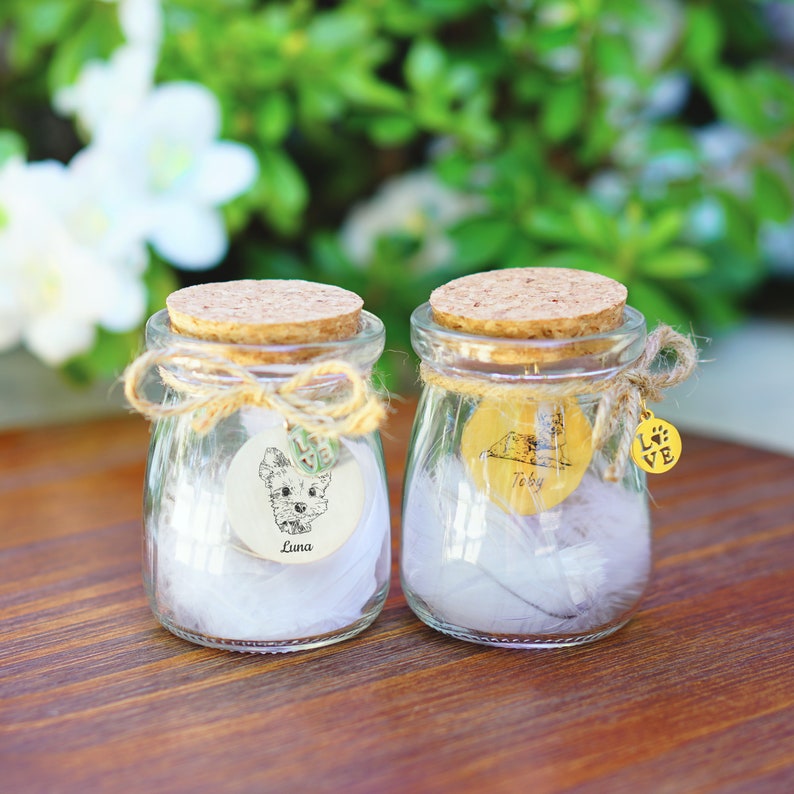 May include: Two small glass jars with cork lids, filled with white feathers and personalized with dog-themed labels. The jars are tied with twine and have a small gold charm with the word "LOVE" and a paw print.