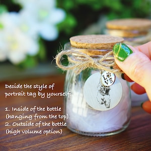 May include: A clear glass bottle with a cork stopper and a twine bow. A silver charm with a paw print and the word "LOVE" hangs from the bottle. The charm also features a black and white silhouette of a dog. The bottle is filled with white tissue paper. The text "Decide the style of portrait tag by yourself: 1. Inside of the bottle (hanging from the top) 2. Outside of the bottle (high volume option)" is visible in the image.