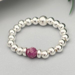 May include: A stretch ring composed of polished silver-toned spherical beads arranged in a circular band. A single, faceted, deep magenta-coloured bead is positioned as a focal point, flanked by two smaller, flat, silver-toned spacer beads.