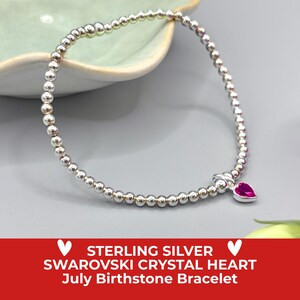 May include: A silver beaded bracelet with a heart-shaped July birthstone charm. The bracelet is displayed on a grey surface with a pink flower and a green leaf in the background. The text on the image reads: STERLING SILVER SWAROVSKI CRYSTAL HEART July Birthstone Bracelet.