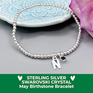 May include: A silver beaded bracelet with a charm featuring the letter "N" and a small green crystal. The bracelet is displayed on a light blue plate and a grey surface, with a pink rose in the background.