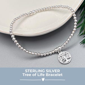 May include: A sterling silver beaded bracelet featuring a Tree of Life charm. The bracelet is presented on a white plate and a grey surface, with green foliage. The text "STERLING SILVER Tree of Life Bracelet" is visible.