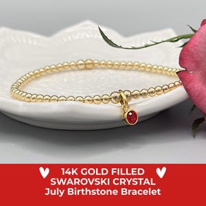 May include: Gold-filled bracelet with small beads and a red Swarovski crystal charm. The bracelet is displayed on a white, decorative plate, with a pink rose in the background. The text on the image reads "14K Gold Filled Swarovski Crystal July Birthstone Bracelet."